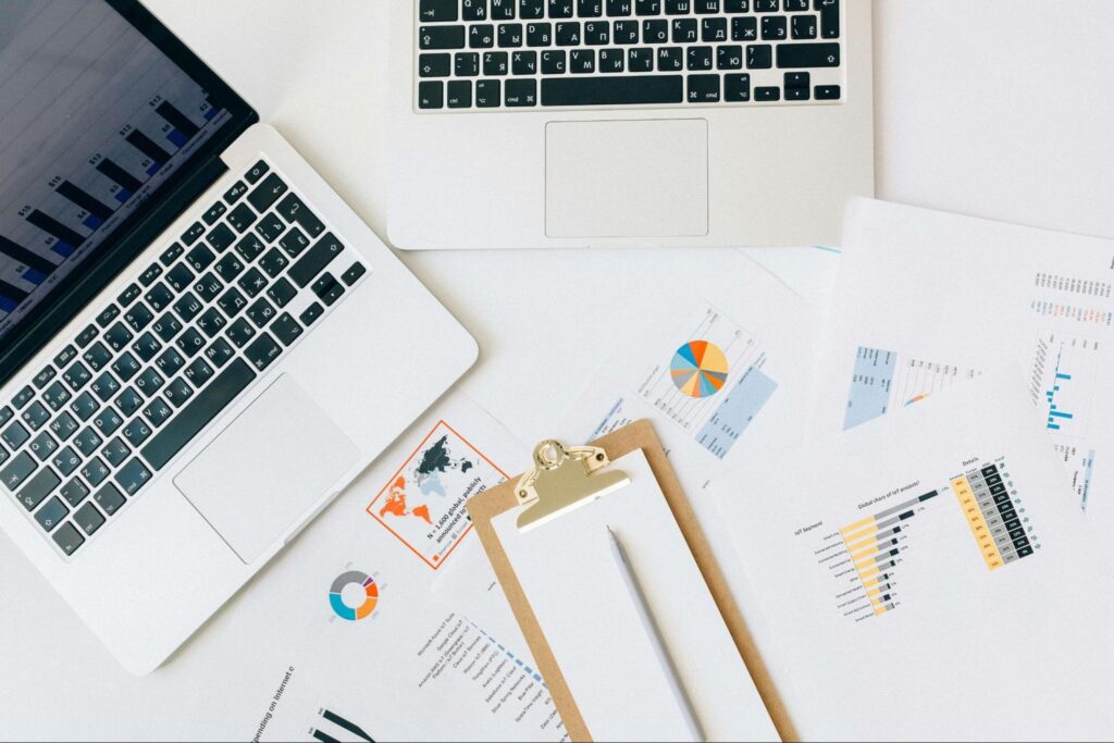 Two laptops and a clipboard shown beside several documents with graphs